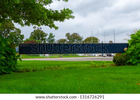 Close view of the entrance sign of park. Located between two shrubs, green grass in the foreground and grey cloudy sky in the background.