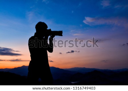 Silhouette of a man who is professional photographer is using a dslr camera at twilight,orange and blue sky on top of the moutain background.Copy space.Ideal for use in documentary, adventure tourism.