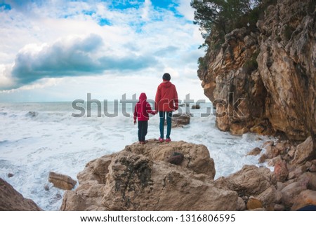 A woman with her son are standing on a stone and looking at the sea. The boy with his mother look at the waves. The child is walking along the ocean shore. Silhouette of mom and child against the sky.