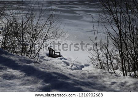Winter scene at a lakeside. Frozen water, partial snow covered, sunny day. A forgotten garden chair standing in deep snow at the lakeside in between bare branches, waiting for the summer. No persons.