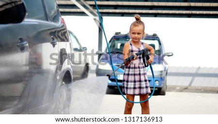 Horizontal image pretty adorable little preschool girl in summer dress helping to parents use coin-operated self-serve car wash, clean auto exterior holding high-pressure sprayer standing outdoors