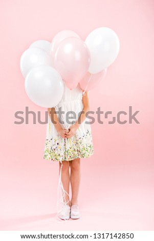 child in delicate white dress hiding behind white and pink air balloons on pink background