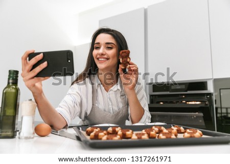 Attractive young woman cooking tasty cookies on a tray while standing at the kitchen, taking a selfie