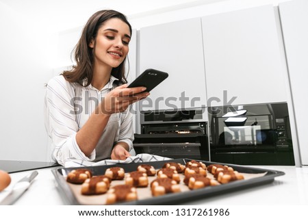 Attractive young woman cooking tasty cookies on a tray while standing at the kitchen, taking a picture