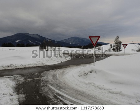 German road with foot high snow and two yield traffic signs on an intersection and mountains in the background with a thick cloudy sky with tire tracks on the road in the snow.