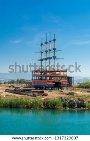 A large sailing ship in a pirate style is moored on the shore for repair against a blue sky. Photos of ship from sea. The concept of summer holidays, sports, tourism 