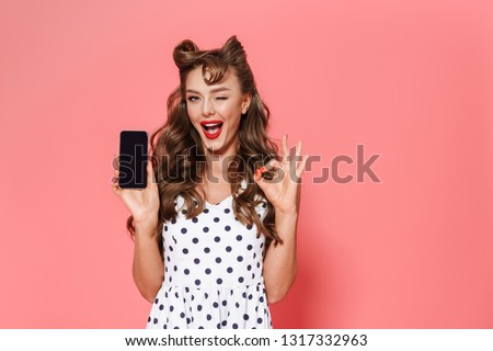 Portrait of a beautiful young pin-up girl wearing dress standing isolated over pink background, showing blank screen mobile phone