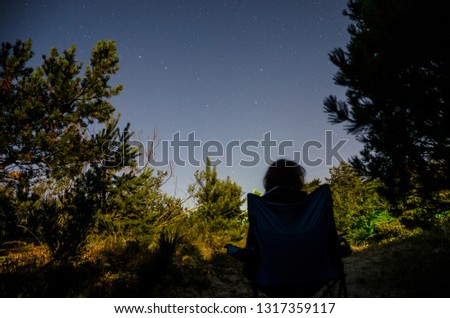 Person sitting in backpacking chair under starry sky between the pines of coastal forest in Latvia, Pape.