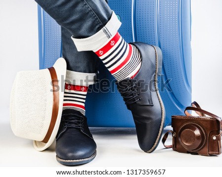 Stylish suitcase, men's legs, vintage camera, sunhat, multicolored socks with a nautical theme and shoes on a white, isolated background. Close-up. Concept of style, fashion, beauty and travel