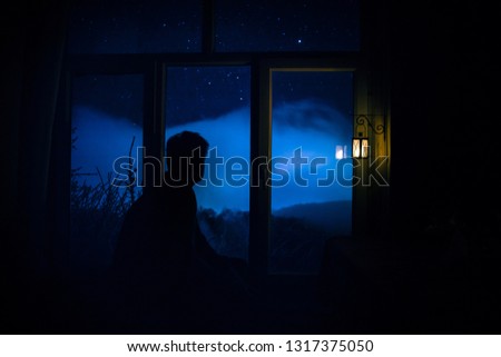 Silhouette of a man looking a dreamlike galaxy through a window. Fantasy picture with old vintage lantern at the window inside dark room.