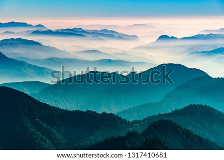 View of Himalayas mountain range with visible silhouettes through the colorful fog from Khalia top trek trail. Khalia top in himalayan region of Kumaon, Uttarakhand, India. Royalty-Free Stock Photo #1317410681