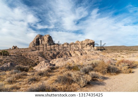 Bright blue sky with white clouds, dirt road and rock formations. Rocky desert landscape with dry brushes and brush.