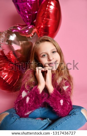 Close-up of cute girl in a studio, smiling widely and playing with pink balloons. She wears sweater and jeans. Folded her hands palms in the shape of a heart.