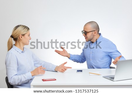 Brutal boss in stylish sunglasses in a blue shirt can not believe his blonde caucasian employee sits by the desk in the office with the white background.