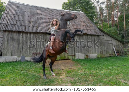 Girl astride horse, who costs on hinder legs, on background of old wooden barn. Riding lessons