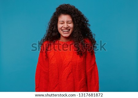 Portrait of happy young cheerful Latin American girl with beautiful black curly hair laughing from funny jokes, has fun, dressed in a red sweater isolated against a blue background
