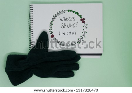 White page of a notebook where is written with nice handwriting: Where is spring,I am cold, isolated on the green background. Hello spring concept.Leaves and flowers as a frame, glove showing a like. 