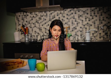 
Peautiful long-haired Asian woman working as a programmer design on a laptop computer in her home kitchen at the dining table with pizza and apples.