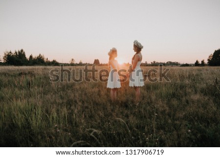 mother and daughter together in white dresses with braids and floral wreaths in boho style in the summer field at sunset. Added a small grain simulation of film photography