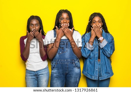 Portrait of three excited african women covering mouthes while telling secrets over yellow background