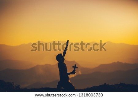 Young man sitting holding christian cross and praying with cry for worshipping God at sunset background. christian silhouette concept.