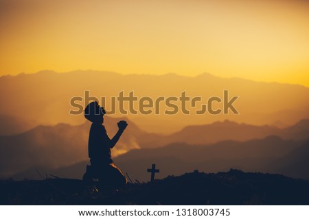 Young man kneeling down and praying with christian cross at sunset background. christian silhouette concept.