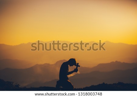 Young man kneeling down praying and holding christian cross for worshipping God at sunset background. christian silhouette concept.