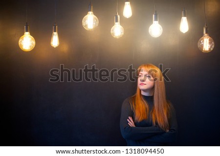 A young woman stands against a black wall under bright lamps. Concept ideas and thinking.
