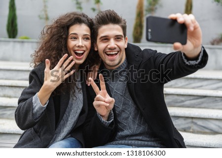 Image of european couple man and woman 20s in warm clothes taking selfie photo on cell phone while sitting on stairs outdoor