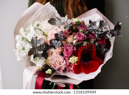 Very nice young woman holding big and beautiful ombre bouquet of fresh roses, eustoma, matthiola, anemone, eucalyptus, flowers in white pink and red colors on the grey wall background 