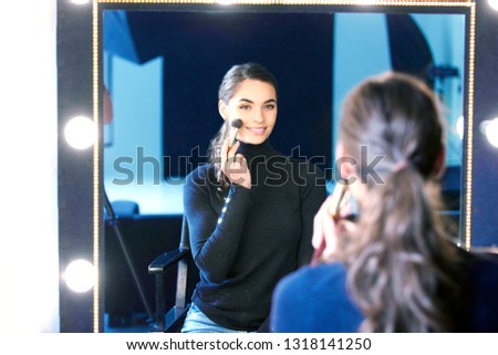 Portrait of gorgeous young woman applying her makeup while sitting at mirror in the photostudio.