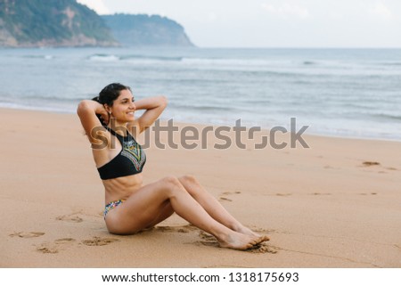 Cheerful fit young woman in bikini doing sit-ups at the beach. Outdoor fitness and healthy lifestyle.