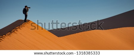 Young Asian man traveler and photographer standing on the top of sand dune photographing sunrise or sunset in desert of Namibia, Africa. Travel photography concept