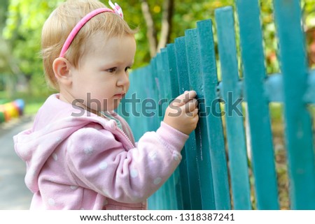 Cute baby girl stay near the fence and focused on something. The child carefully paints the chalk. Paint a green fence. Walking with the baby in the summer or in spring. The child will know the world.