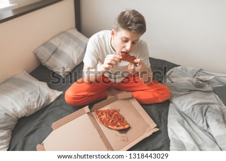 Happy young man sitting at home on a bed with a box of pizza, bites a piece of pizza with his eyes closed and gets pleasure. Giy is home to the bed and eats a delicious pizza.