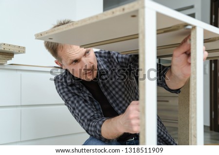 The man is assembling the furniture at home. He is mounting and fastening the frame of the chest of drawers.