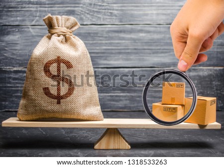 Magnifying glass is looking at the bunch of boxes. bag of money on the scales. Conceptual trade balance between countries and unions, trade and exchange of goods. Economic relations between subjects Royalty-Free Stock Photo #1318533263