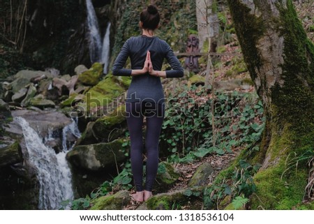Young slim woman practicing yoga outdoors in moss forest on background of waterfall. Unity with nature concept. Girl standing with prayer hands.