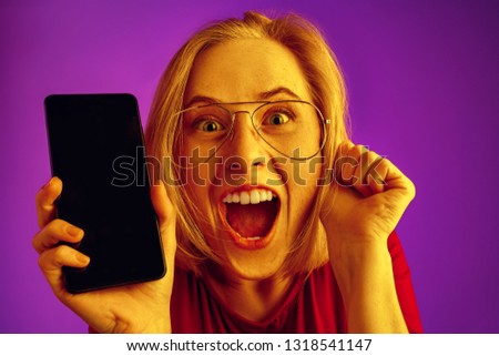Portrait of a confident casual girl showing blank screen of mobile phone over neon studio background.