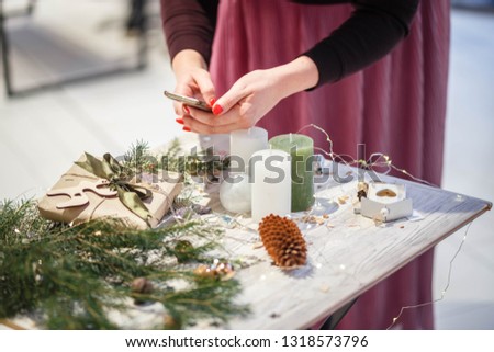 Female hands taking pictures of holiday decorations. Christmas candles, garland, branch of pine, baubles and amethyst pendants on the white wooden table. Celebration and decoration concept. 