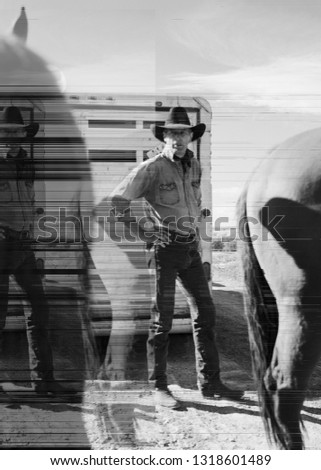 Mid-adult cowboy standing with his hand on his hip next to horses on a ranch.