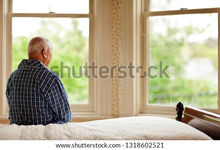 Senior man looking out his bedroom window while sitting on a bed in his pajamas.