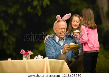 Senior man wears a bunny ear headband and smiles as he sits and takes afternoon tea with two young girls.