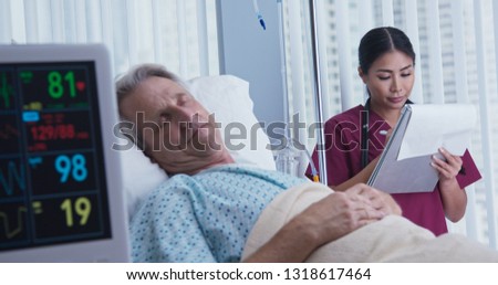 Nurse taking notes on chart with senior Caucasian male patient recovering in hospital bed in foreground. Japanese woman doctor monitoring recovery