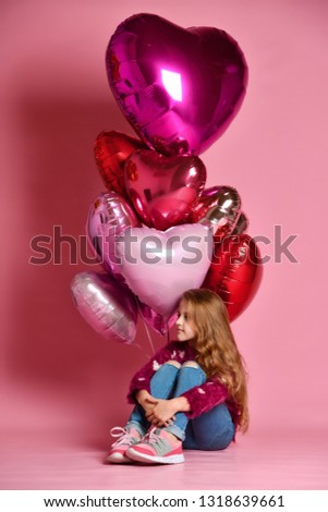 A little 7 year old girl is sitting on the floor holding a bunch of pink heart-shaped balloons. Celebrates birthday and Valentine's Day. Hangout. Birthday party.