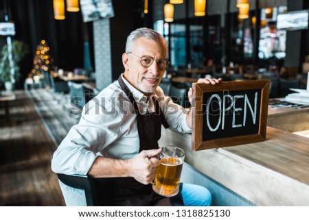 smiling owner of pub holding open sign and glass of beer at bar counter