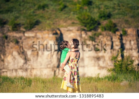 Mother kisses the boy. A woman is walking with her son across the field. The child is sitting at mother's arms and hugs her. Mom's embrace.