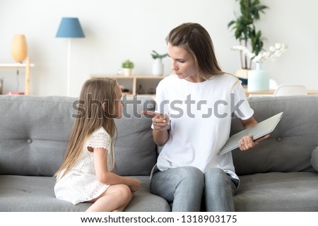 Strict mother scolding little daughter for long laptop use, angry woman with preschool girl sitting on couch in living room, holding computer in hands, parent reprimanding, lecturing child Royalty-Free Stock Photo #1318903175