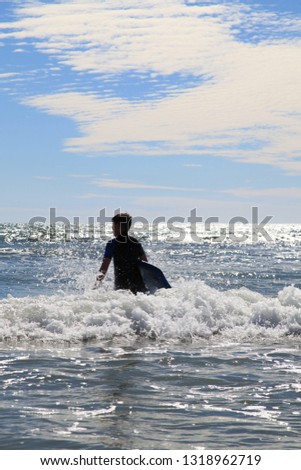Boy with surf board walking into sea, against a blue slightly cloudy sky