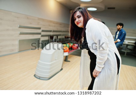 South asian woman standing at bowling alley with ball on hands. Girl is preparing for a throw.